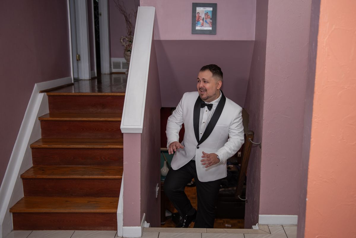 A man in a white tuxedo descends a wooden staircase, smiling and gesturing, at a warmly lit indoor event.