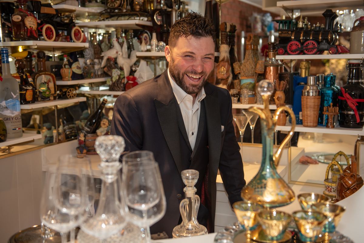 A man in a tuxedo smiles behind a counter filled with glassware and decanters in an upscale setting.