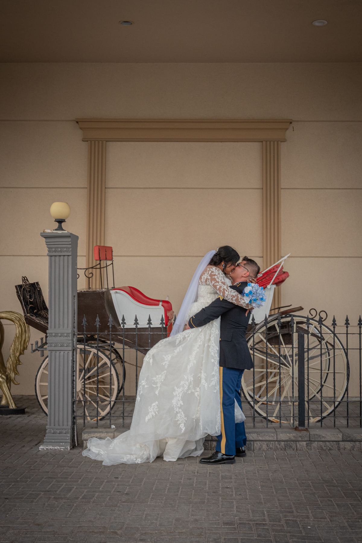 A bride in a lace gown and veil kisses her groom in military uniform by a vintage carriage, creating an intimate moment.