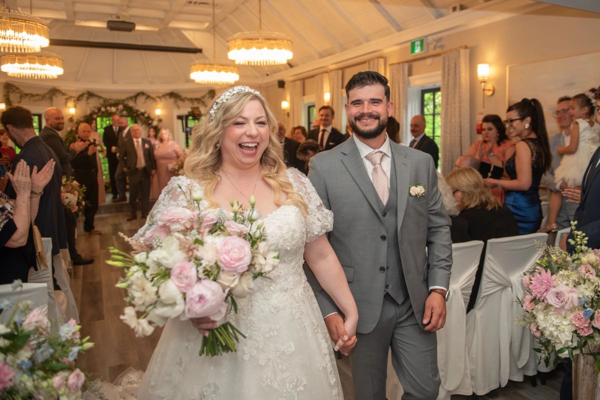 A smiling bride and groom walk down the aisle, hand-in-hand, as guests applaud under elegant chandeliers.