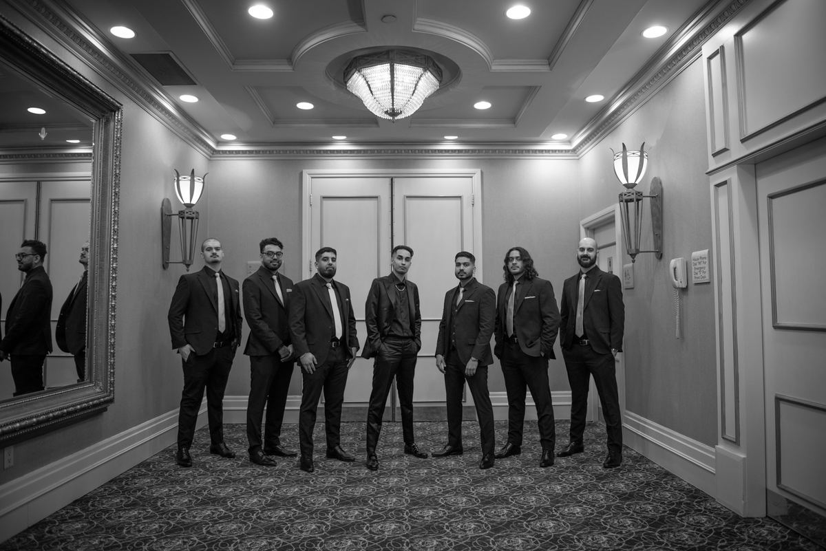 Seven formally dressed men stand in an ornate hallway under soft lighting, their expressions serious.