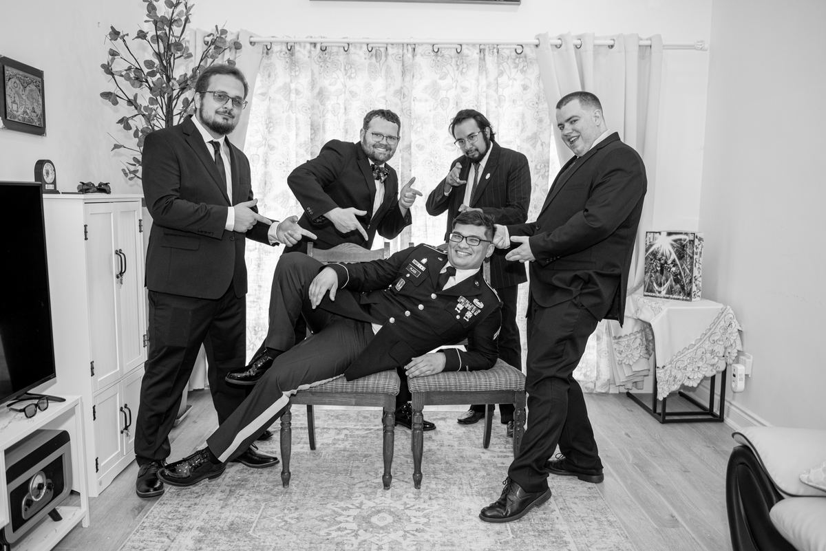A groom in military uniform relaxes as four smiling groomsmen pose playfully around him in a cozy room.