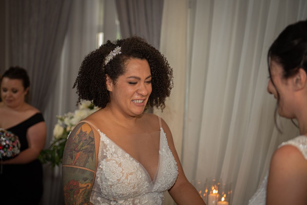A bride with curly hair and a tattooed arm smiles warmly at a bridesmaid near soft candlelight and white curtains.