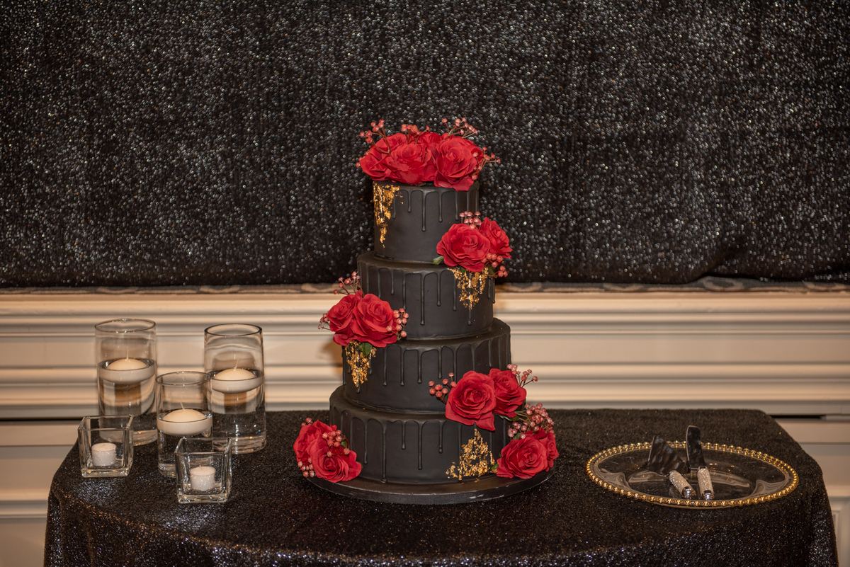 A black drip cake with red roses and gold accents sits on a glittering tablecloth near candles and tools.
