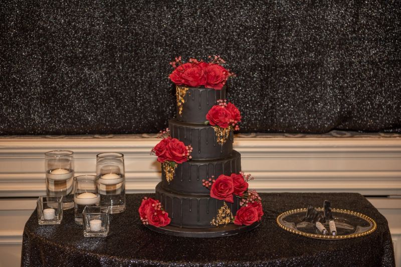 A black drip cake with red roses and gold accents sits on a glittering tablecloth near candles and tools.