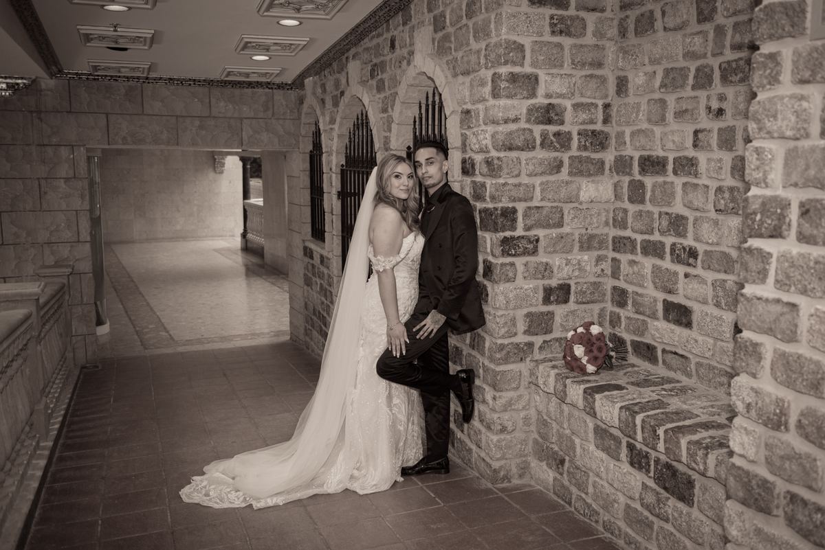 A newlywed couple in a stone-walled corridor, the bride in white leaning against her groom with one leg up on the wall.