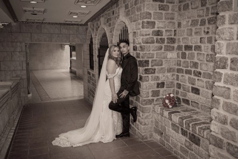 A newlywed couple in a stone-walled corridor, the bride in white leaning against her groom with one leg up on the wall.