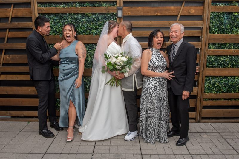 The bride and groom kiss joyfully while surrounded by laughing family members near a wooden fence with greenery.