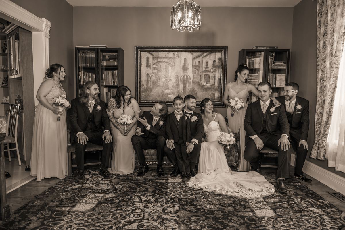 Bride and groom surrounded by laughing wedding party, sitting on patterned rug near bookshelves and painting.
