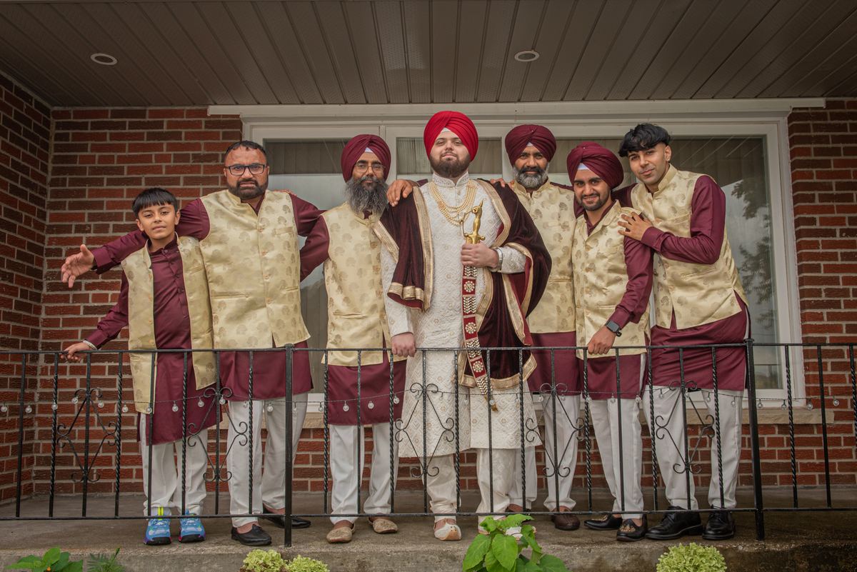 A groom in white with a red turban and sword stands with his groomsmen and a young boy in matching maroon and gold attire.