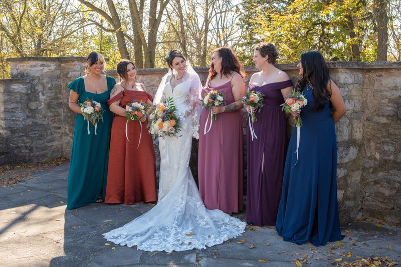 A bride and five bridesmaids in colorful gowns stand outdoors beside a stone wall, surrounded by fall foliage.