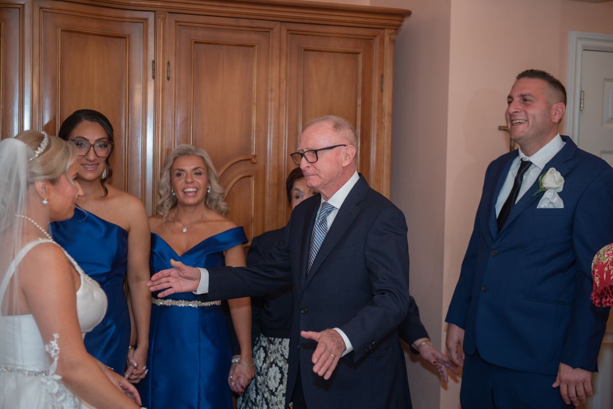 An older man in a suit gestures warmly to the bride and smiling bridesmaids indoors near wooden cabinets.