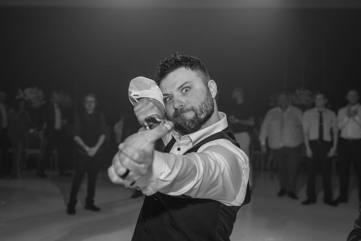 A groom in formal wear pops open a champagne bottle with an intense smile, surrounded by blurred guests.