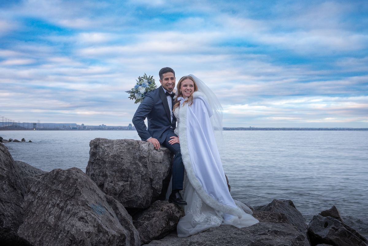 A newlywed couple poses joyfully on rocks by a waterfront, smiling at the camera as a cityscape looms in the distance.