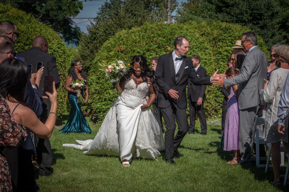 A joyful bride in a white lace gown, assisted by her groom, walks down a grassy aisle lined with applauding guests.
