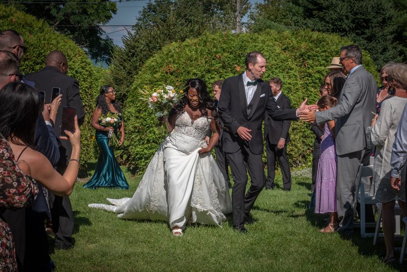 A joyful bride in a white lace gown, assisted by her groom, walks down a grassy aisle lined with applauding guests.