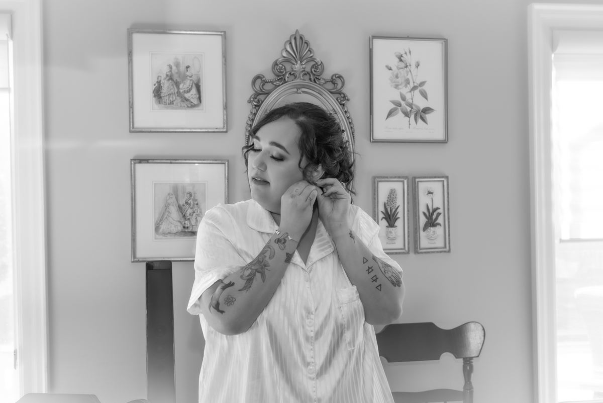 A bride adjusts her earring in front of ornate framed prints, with soft natural light illuminating the room.