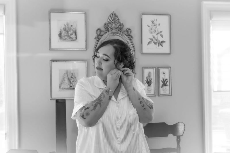 A bride adjusts her earring in front of ornate framed prints, with soft natural light illuminating the room.