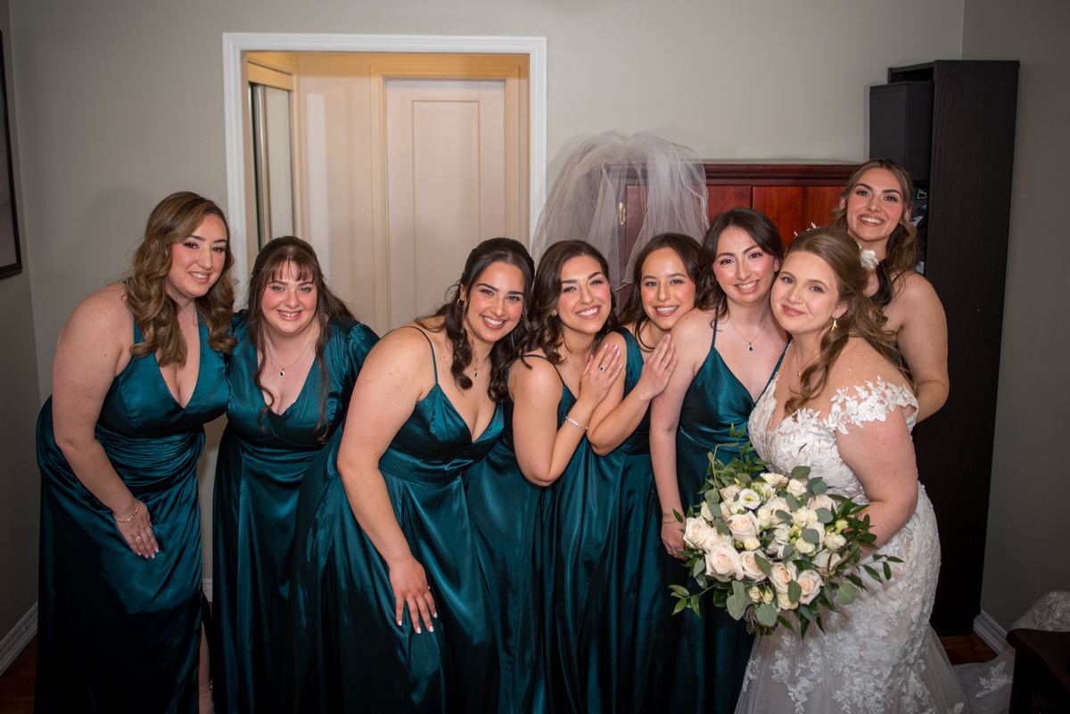 A bride in a white lace gown with her bridesmaids in teal satin gowns smiles for a group portrait near a draped veil.