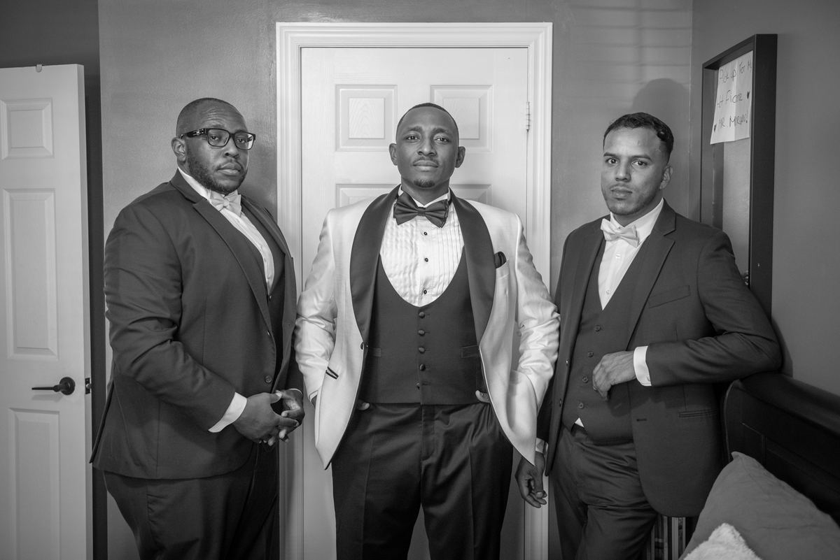 Three formally dressed men stand together in a softly lit room, with the center figure in a white tuxedo jacket.