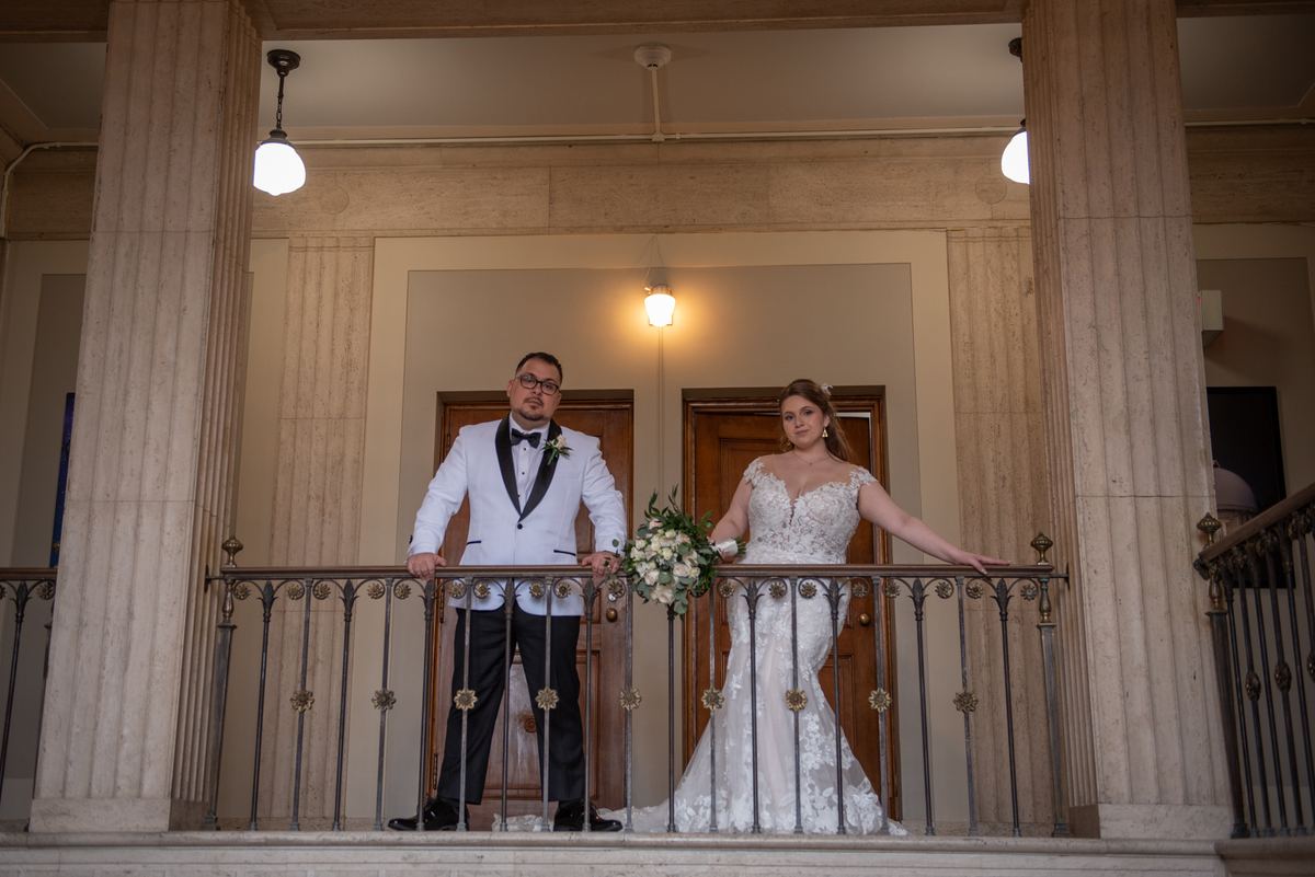 A bride and groom stand on an ornate balcony inside a grand hall, leaning on the railing with the bride holding a bouquet.