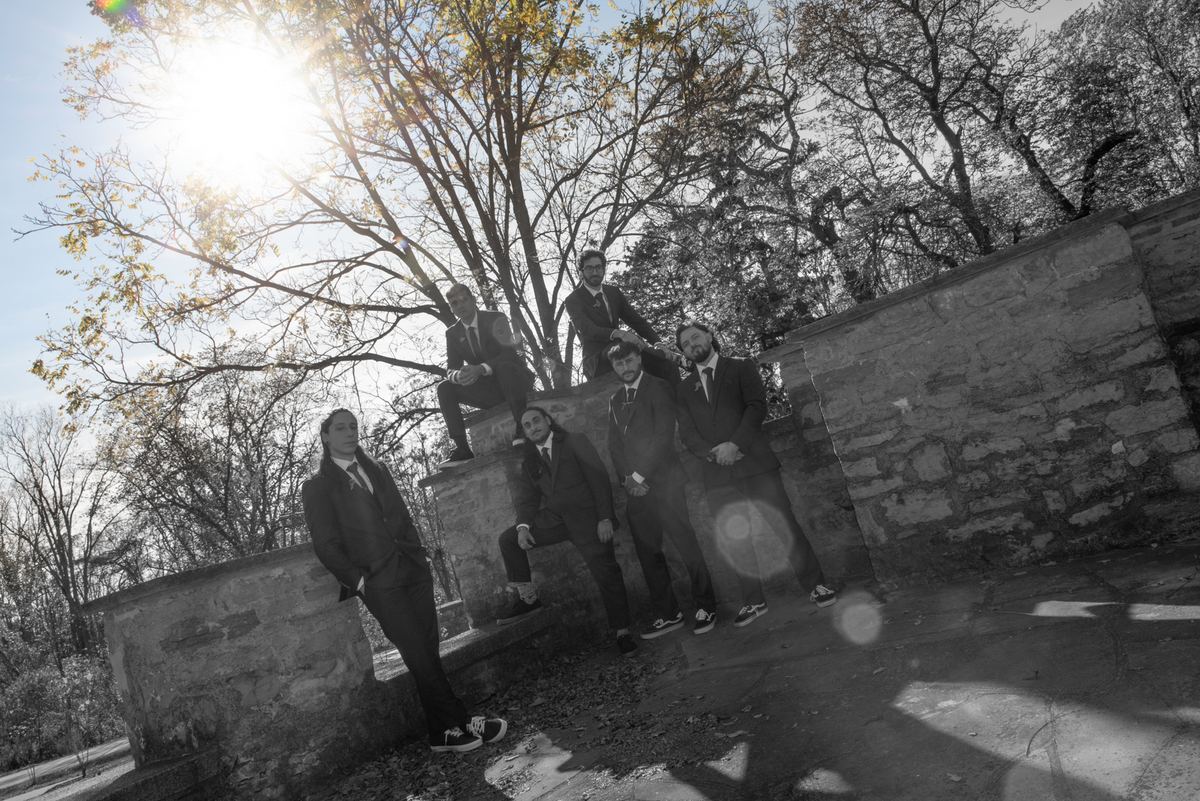 Six men in suits pose casually outdoors on stone steps and walls, with sunlight casting long shadows.