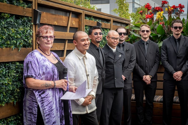 A man in a white barong tagalog holds papers while standing with others beside a wooden fence and greenery.