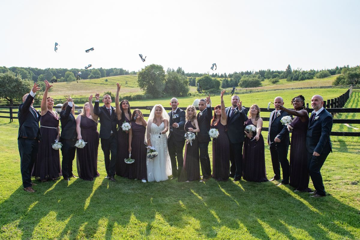 Stephanie and Carmelo's wedding party in a sunlit field, arms raised as they toss flower petals into the air.
