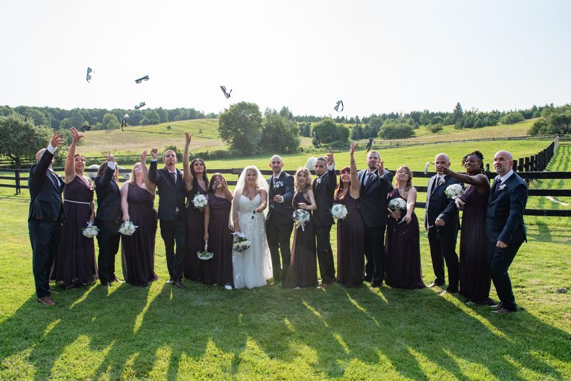 Stephanie and Carmelo's wedding party in a sunlit field, arms raised as they toss flower petals into the air.