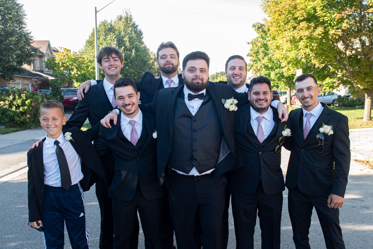 Groomsmen and a young boy in formal suits pose joyfully outdoors on a sunny afternoon, arms around each other.