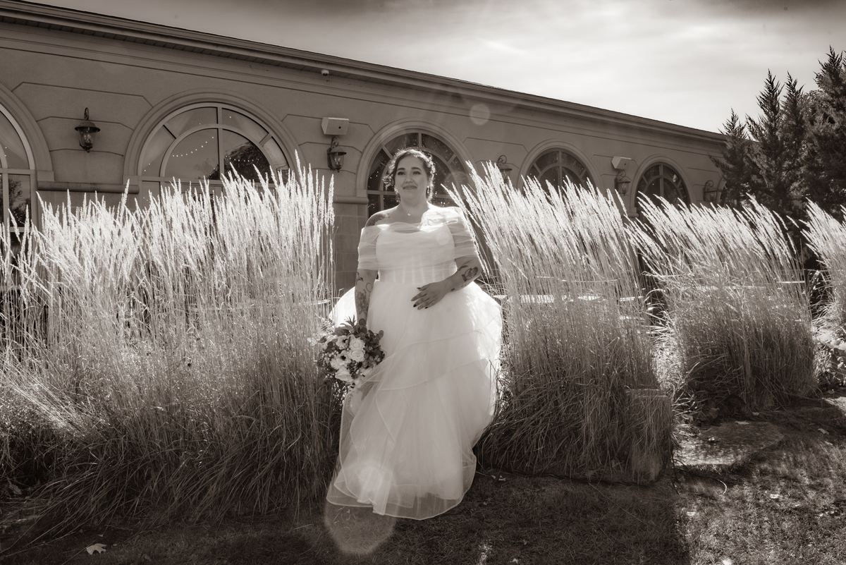 A bride in a flowing gown stands among tall grasses, smiling at the camera with an arched building behind her.