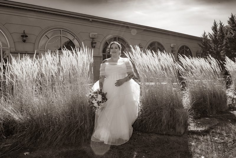 A bride in a flowing gown stands among tall grasses, smiling at the camera with an arched building behind her.