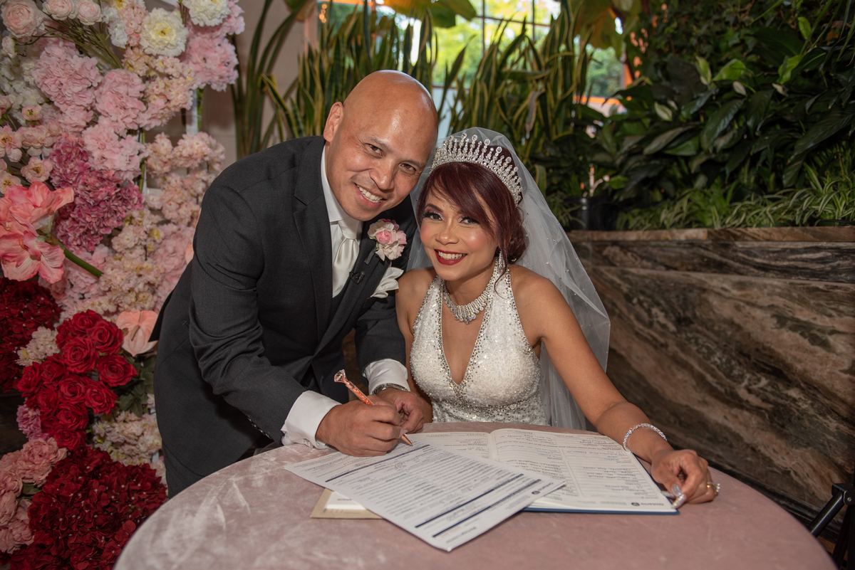 A smiling bride in a white gown and tiara and her dark-suited groom sign marriage documents surrounded by flowers.