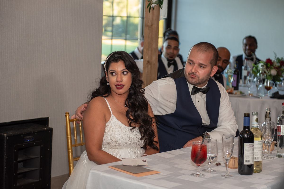 A bride and groom sit at a table, both looking toward the side with playful expressions, his hand on her shoulder.