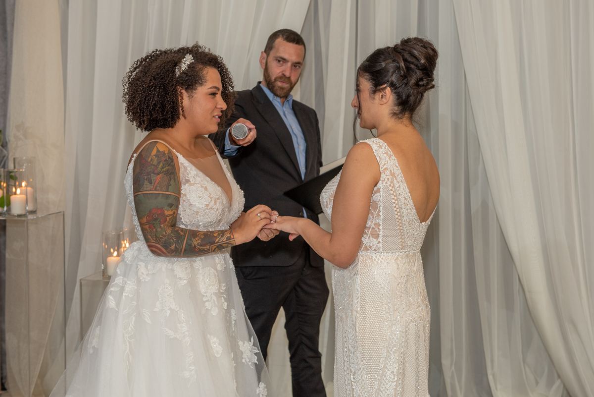 Two brides exchange rings under candlelight, one in a lace gown, the other in a sleeveless dress with an open back.
