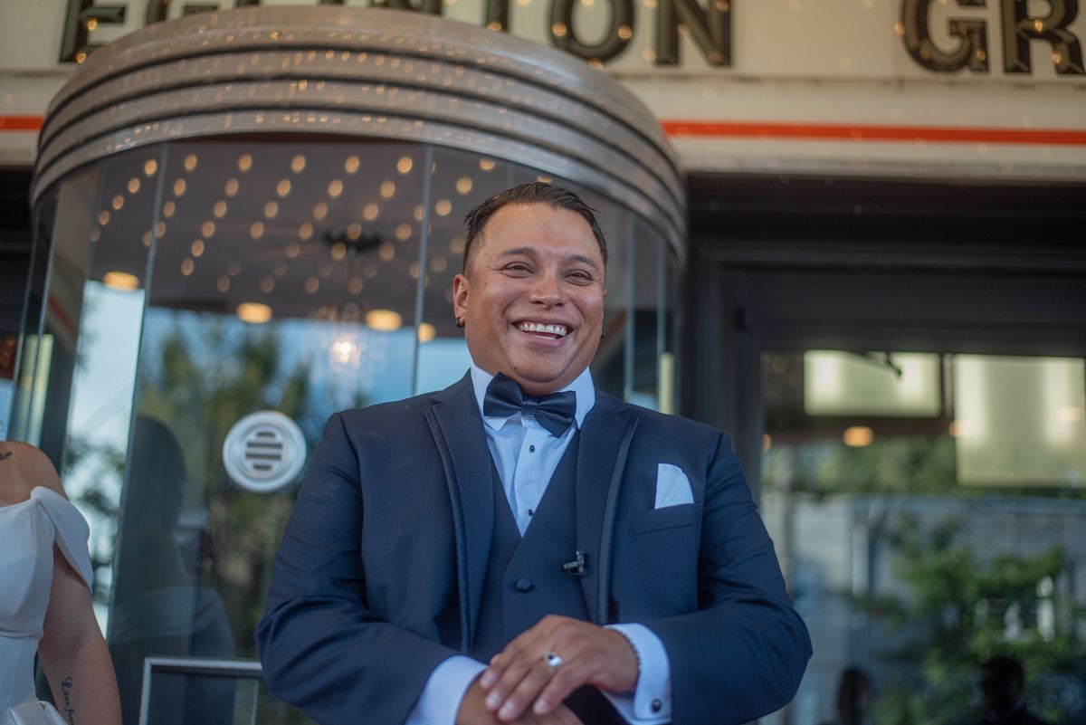 A groom in a navy tuxedo smiles next to a modern building with a revolving door and blurred windows.