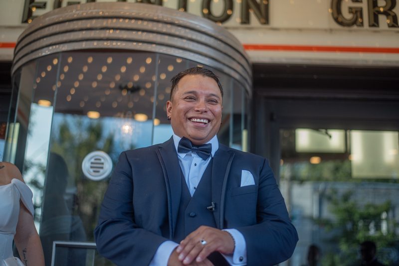 A groom in a navy tuxedo smiles next to a modern building with a revolving door and blurred windows.