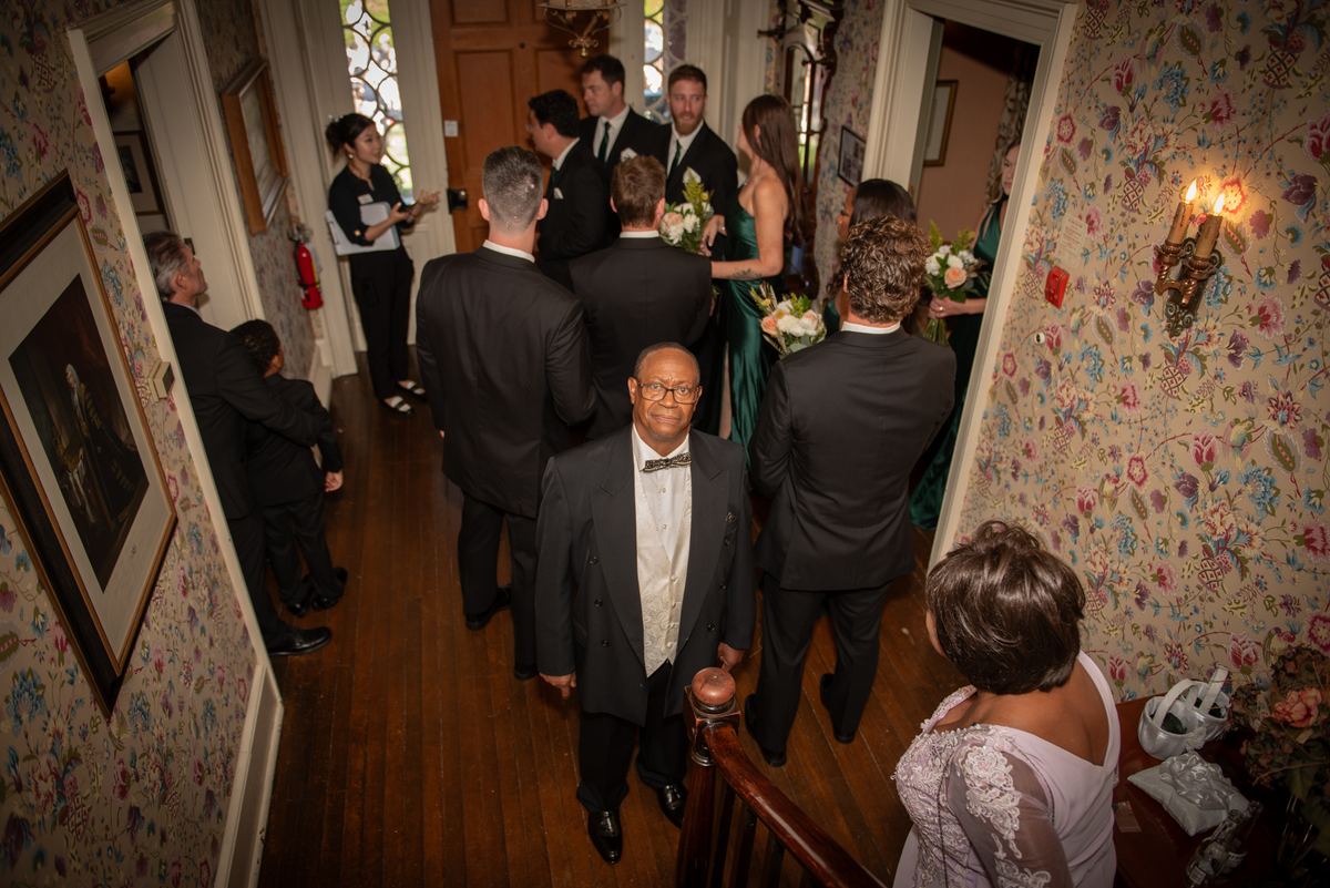 A wedding procession moves down an ornately wallpapered hallway with guests in formal attire following the bride.