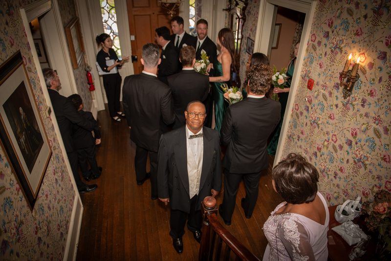 A wedding procession moves down an ornately wallpapered hallway with guests in formal attire following the bride.