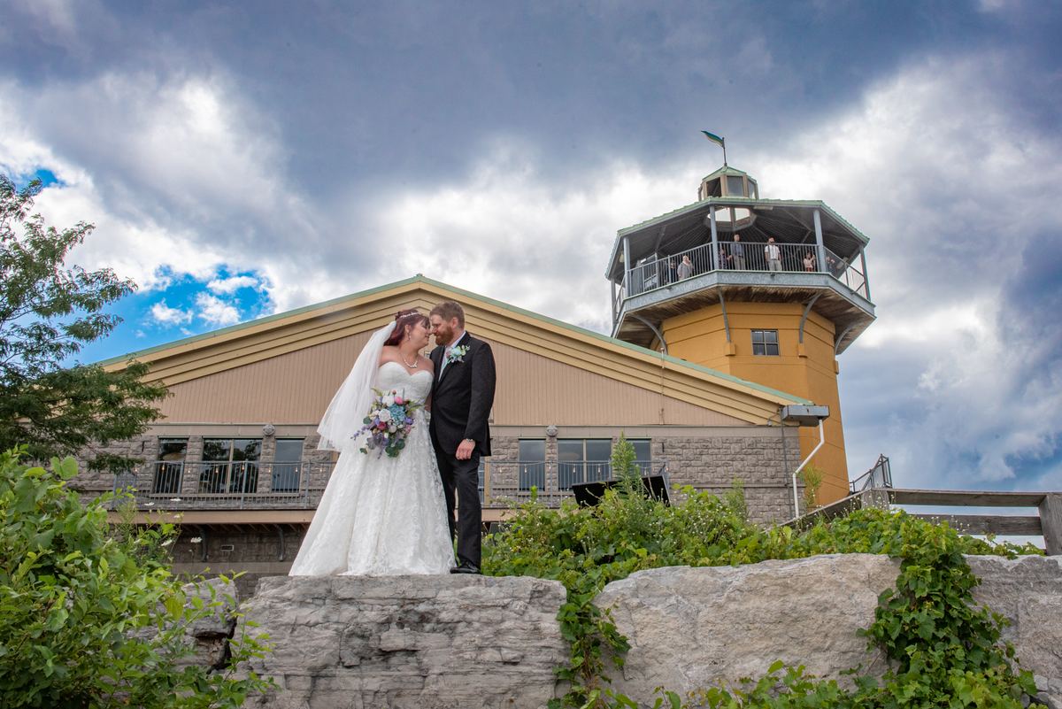 A bride and groom kiss on a stone ledge under a partly cloudy sky, with a yellow tower visible behind them.