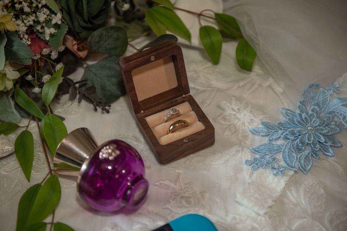 Wedding rings rest in an open wooden box surrounded by lace and greenery, suggesting wedding preparations.