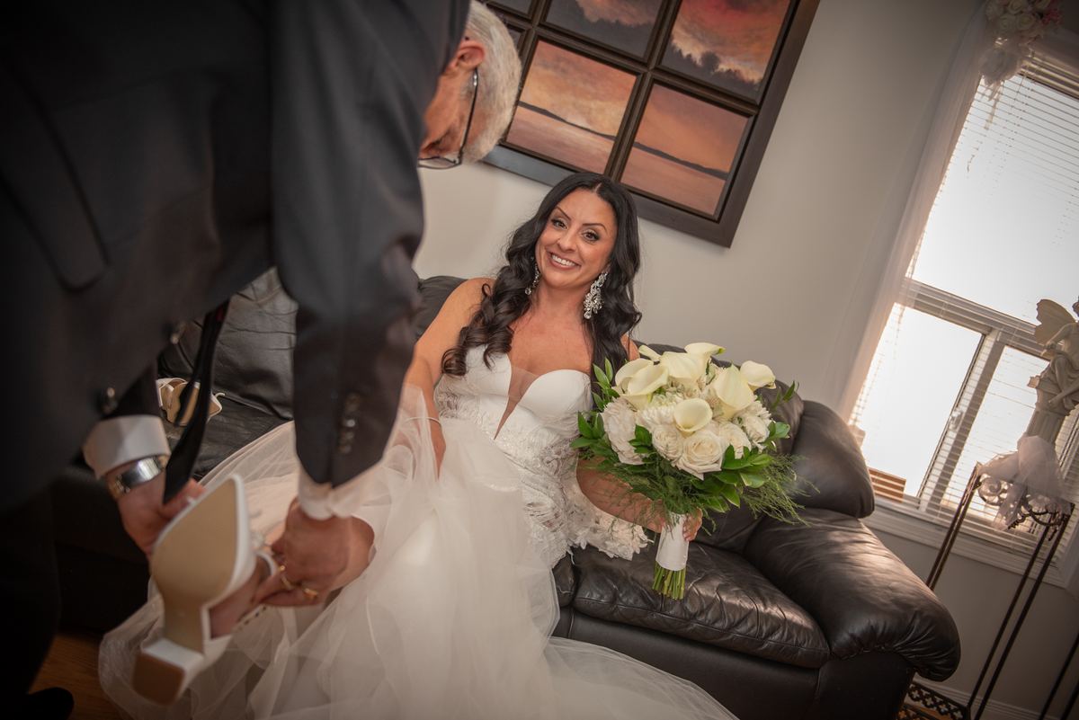 A bride sits smiling as her father helps her into her wedding shoes, illuminated by soft natural light.