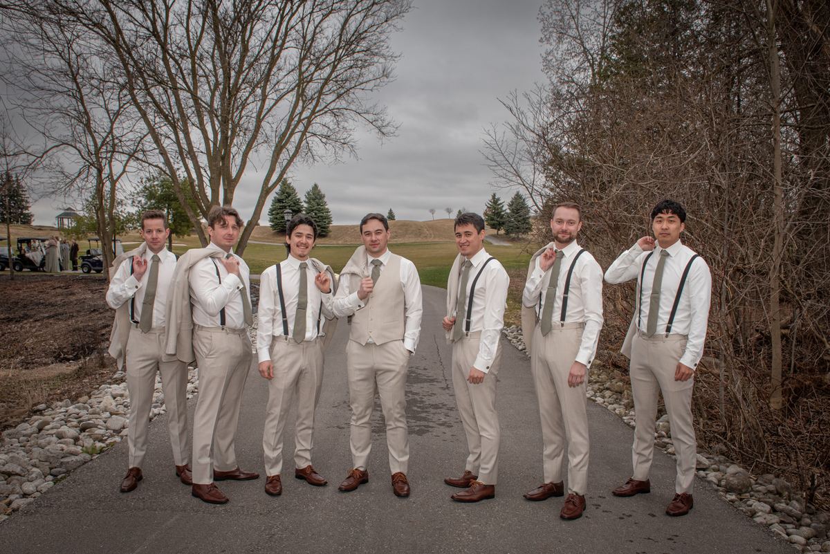 Seven groomsmen in beige suits stand outdoors with cheerful expressions during a pre-wedding photoshoot.