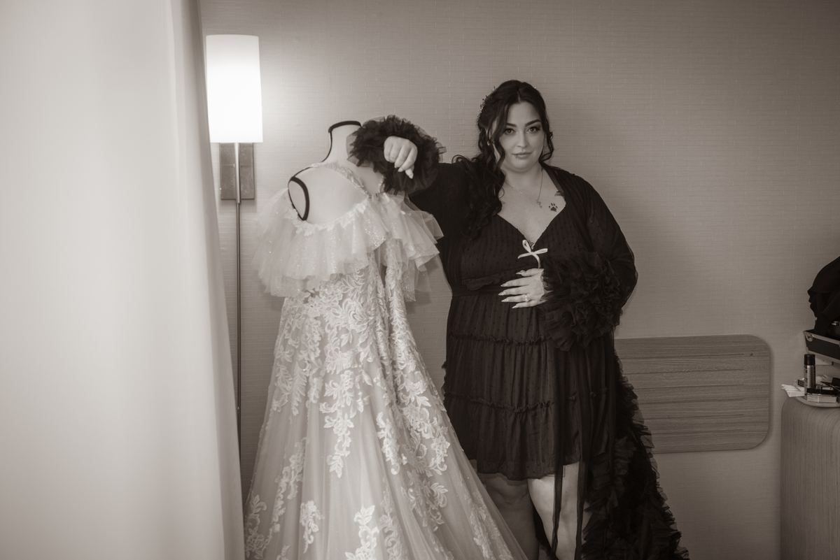 A woman stands beside a mannequin draped in an elaborate wedding gown, gently adjusting its feathered collar.