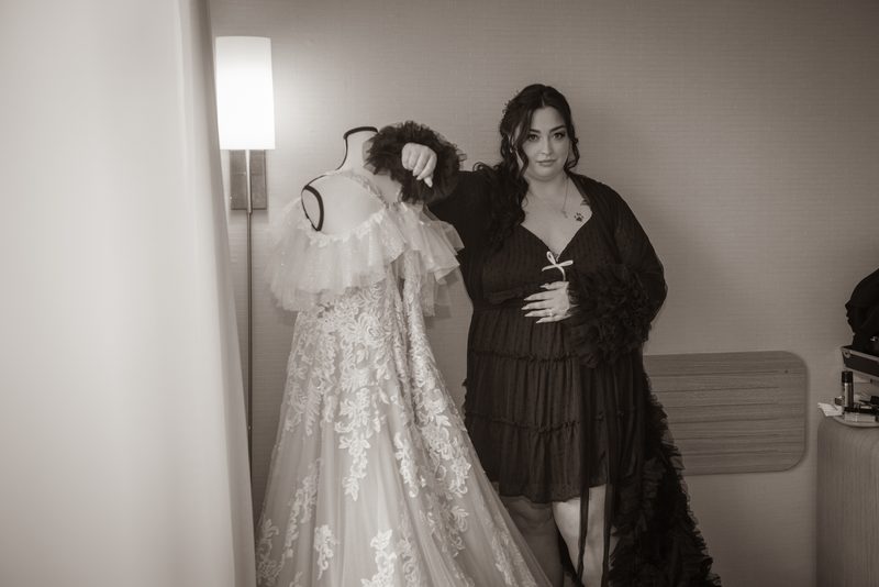 A woman stands beside a mannequin draped in an elaborate wedding gown, gently adjusting its feathered collar.
