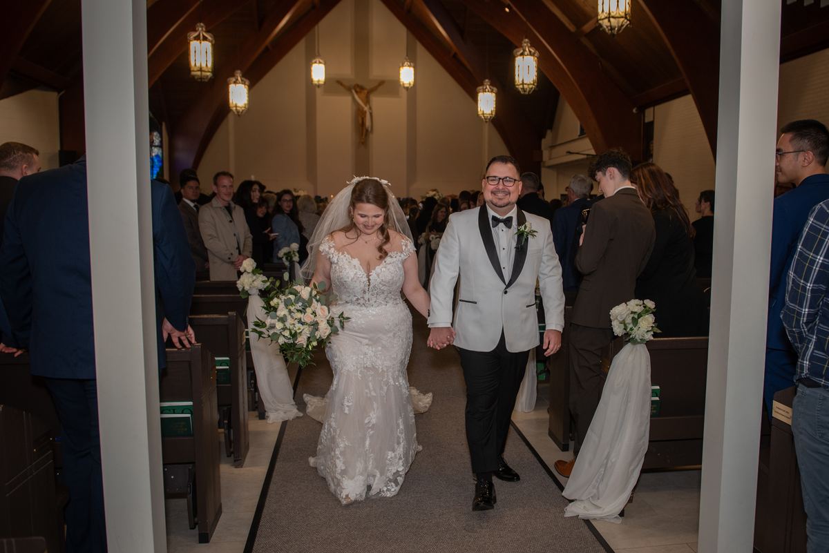 A bride in a lace gown and veil walks down the aisle with a groom in a white tuxedo, illuminated by hanging lanterns.