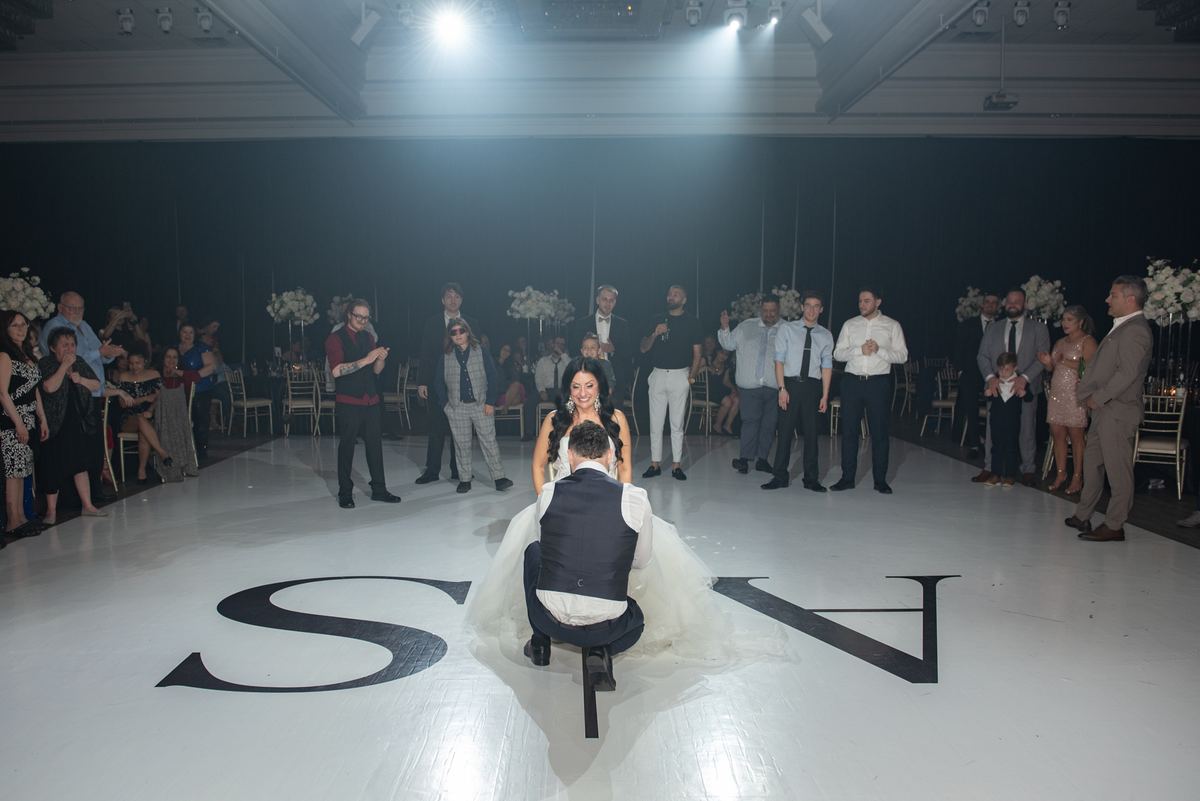 A groom kneels on a white dance floor marked with initials as guests in formal attire clap and smile.