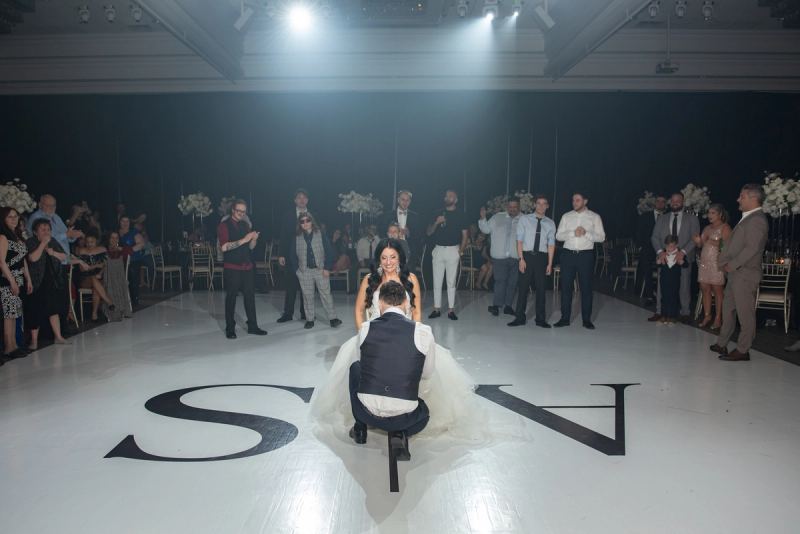 A groom kneels on a white dance floor marked with initials as guests in formal attire clap and smile.
