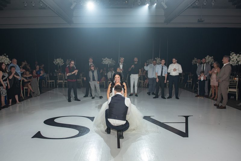 A groom kneels on a white dance floor marked with initials as guests in formal attire clap and smile.
