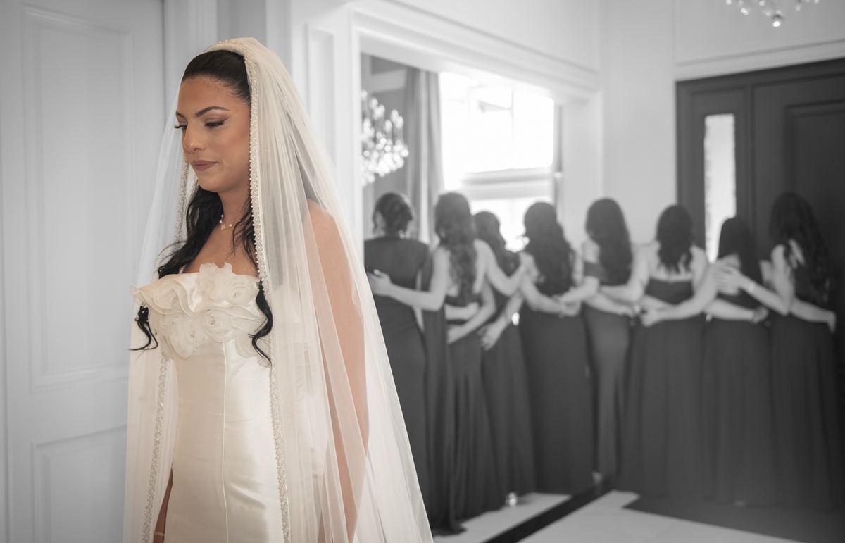Bride in white gown and veil stands contemplatively as bridesmaids wait behind her in an elegant room.